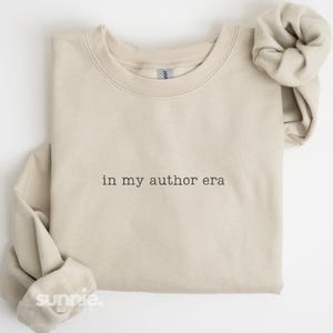 May include: A beige crewneck sweatshirt featuring the black embroidered text "in my author era" centered on the chest. The garment has long sleeves with ribbed cuffs and a ribbed neckline, showcasing a clean, simple design with a classic fit.