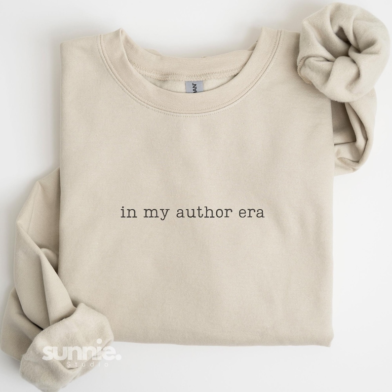 May include: A beige crewneck sweatshirt featuring the black embroidered text "in my author era" centered on the chest. The garment has long sleeves with ribbed cuffs and a ribbed neckline, showcasing a clean, simple design with a classic fit.