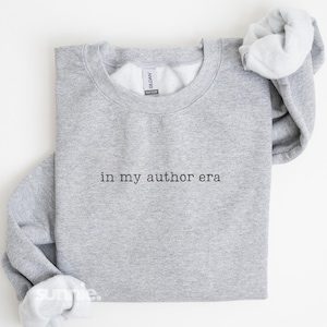 May include: A heather gray crewneck sweatshirt featuring the black text "in my author era" embroidered on the center chest. The garment has long sleeves with ribbed cuffs and a ribbed neckline, with a Gildan brand label visible inside the collar.