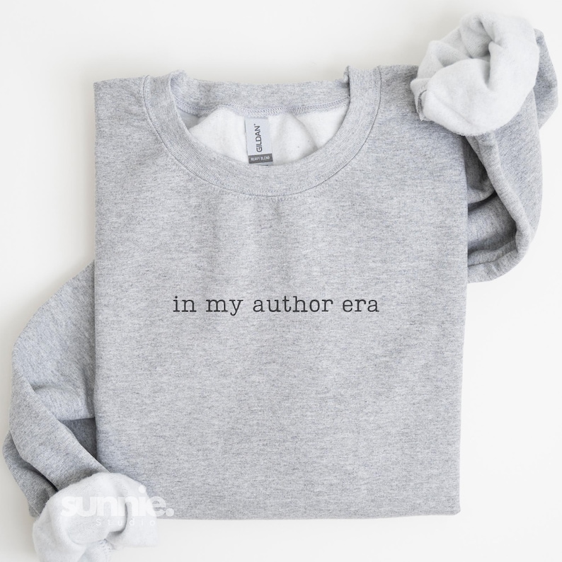 May include: A heather gray crewneck sweatshirt featuring the black text "in my author era" embroidered on the center chest. The garment has long sleeves with ribbed cuffs and a ribbed neckline, with a Gildan brand label visible inside the collar.