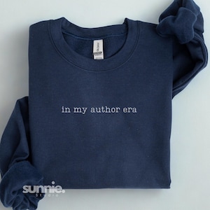 May include: A navy blue crewneck sweatshirt featuring the white embroidered text "in my author era" centered on the chest. The garment has a ribbed collar, long sleeves, and a Gildan brand label visible at the inner neckline. The fabric is solid.