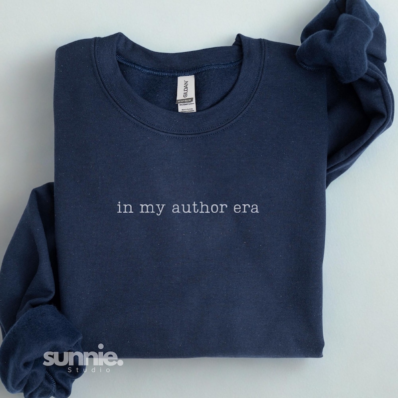 May include: A navy blue crewneck sweatshirt featuring the white embroidered text "in my author era" centered on the chest. The garment has a ribbed collar, long sleeves, and a Gildan brand label visible at the inner neckline. The fabric is solid.