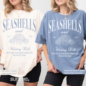 May include: Two t-shirts, one cream and one blue, with the text "SEASHELLS and Wedding Bells" and the year 2026. The shirts feature a seashell graphic and the text "OLIVIA'S BACHELORETTE FRANCE 2026".