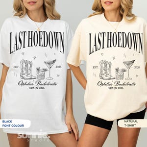 Könnte beinhalten: Zwei kurzärmelige T-Shirts, eines in Weiß und eines in Naturfarben, mit schwarzem Grafiktext LAST HOEDOWN, EST. 2026 und Ophelia's Bachelorette SPAIN 2026. Das Design enthält Illustrationen von Cowboystiefeln und Cocktailgläsern.