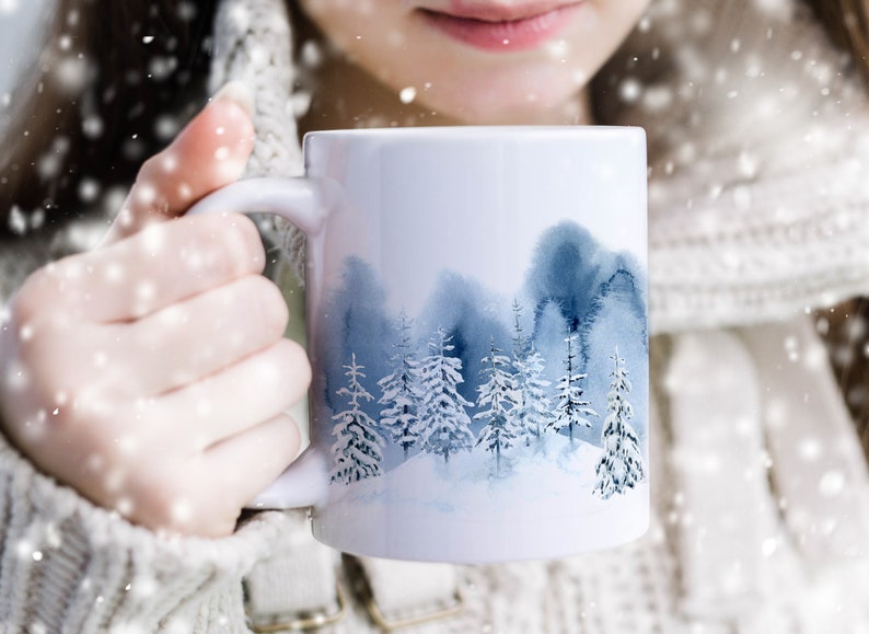 May include: White ceramic mug with a watercolor design of a snowy forest scene. The mug has a blue and white color scheme and features a variety of trees.