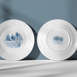 May include: Two white ceramic plates with a blue watercolor design of a winter scene with trees and a small house. The plates are shown from the front and back.