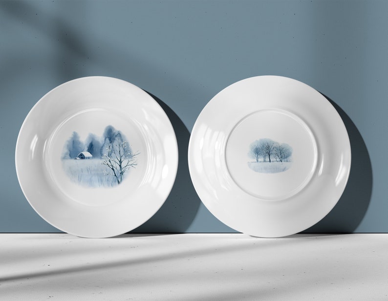May include: Two white ceramic plates with a blue watercolor design of a winter scene with trees and a small house. The plates are shown from the front and back.