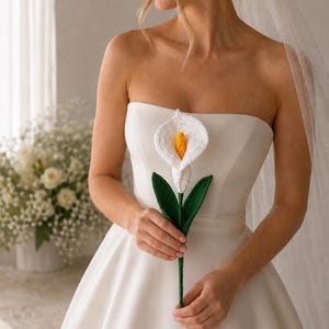 Puede incluir: Vestido de novia blanco sin tirantes con una cala de ganchillo. El lirio tiene una flor blanca con centro amarillo, hojas verdes y tallo verde. La novia lleva velo.