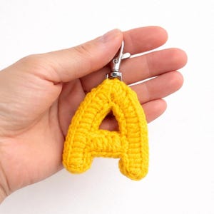May include: A bright yellow crocheted letter "A" keychain. The letter is made of thick yarn and has a silver-colored metal clasp. The keychain is held against a white background.