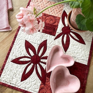 May include: A white quilted table runner with a red and pink floral design. The runner features a pink flower with green leaves, a red and pink heart-shaped bowl, and a red and white geometric pattern.