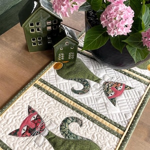 May include: A quilted table runner with a cat design. The runner has a white background with green and yellow accents. The cat's face is red with green floral details. Ceramic houses and pink flowers are in the background, adding a decorative touch.