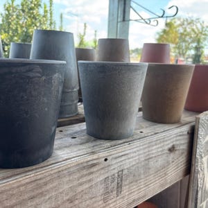 May include: A collection of various sized, gray and brown, tapered plant pots. The pots are arranged on a weathered wooden shelf. The pots have a textured surface and are made of a durable material. The pots are suitable for indoor or outdoor use.