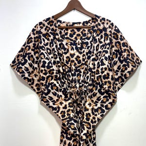 May include: A leopard print dress with a V-neck and short sleeves hangs on a wooden hanger. The dress features a brown, black, and cream-colored animal print. The dress has a drawstring waist and a flowing, floor-length design.