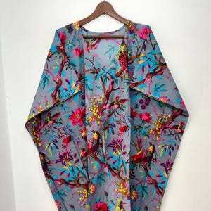 May include: Gray, open-front kaftan with a vibrant floral and bird print. The design features pink flowers, yellow accents, and teal leaves against a gray background. The kaftan is hanging on a wooden hanger.
