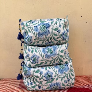 May include: A stack of three rectangular fabric pouches featuring a white base with a blue and green floral print. Each pouch has a quilted texture and a side zipper closure adorned with a small blue tassel featuring gold thread accents.