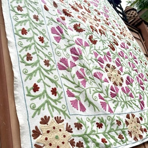 May include: A white cotton fabric with intricate embroidery featuring a floral pattern in shades of pink, green, and brown. The embroidery is done in a traditional style with delicate stitches and a variety of floral motifs.