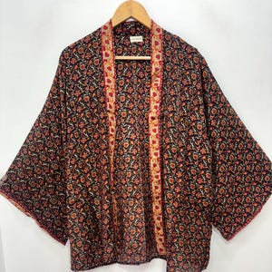 May include: A black kimono with a floral pattern in shades of orange and red. The kimono has wide sleeves and a contrasting trim along the edges. It is hanging on a wooden hanger.