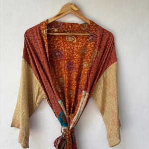 May include: A long-sleeved robe featuring a patchwork design with a mix of orange, rust, and beige tones. The fabric displays intricate paisley and floral patterns throughout, with a tie-waist closure and an open front hanging on a wooden hanger.