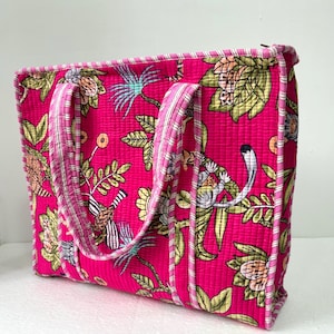 May include: A quilted tote bag with a vibrant pink background and a botanical print featuring flowers, leaves, and birds. The bag has matching pink handles and trim with a striped pattern. The bag is rectangular and has a structured shape.