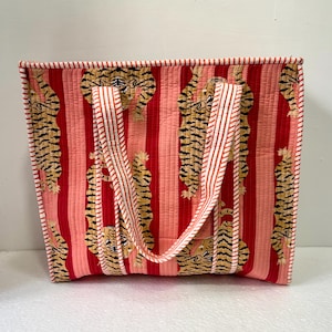 May include: A quilted tote bag with a repeating tiger pattern on a red and pink striped background. The bag has white and orange striped handles and trim. The bag is rectangular and appears to be made of fabric.
