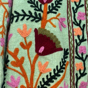 May include: Close-up of a textile with embroidered floral patterns. The design features orange, pink, and burgundy flowers with black and green leaves on a light green background. The embroidery is detailed and textured.