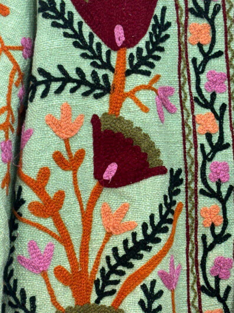 May include: Close-up of a textile with embroidered floral patterns. The design features orange, pink, and burgundy flowers with black and green leaves on a light green background. The embroidery is detailed and textured.