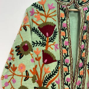 May include: A light green jacket with intricate floral embroidery. The embroidery features red, orange, pink, and green colors. The jacket has a high collar and long sleeves, with the floral design covering the front and sleeves.