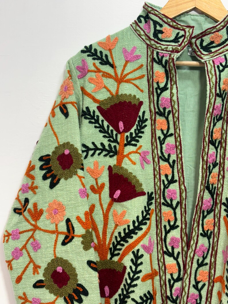 May include: A light green jacket with intricate floral embroidery. The embroidery features red, orange, pink, and green colors. The jacket has a high collar and long sleeves, with the floral design covering the front and sleeves.