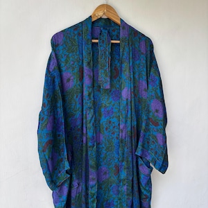 Könnte beinhalten: Ein langärmeliger Morgenmantel mit einem lebendigen Blumenmuster in den Farben Dunkelblau, leuchtendes Türkis und Violett. Das Kleidungsstück hat ein offenes Vorderteil mit passendem Bindegürtel und hängt an einem Holzkleiderbügel an einer Wand.