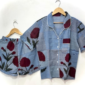 May include: Blue and white patterned pajama set with a floral design. The set includes a short-sleeved button-down shirt and matching shorts. The shirt has a white collar and pocket trim. The shorts and shirt feature a repeating pattern of dark red flowers.