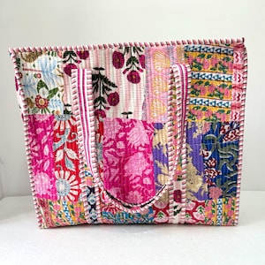 May include: A patchwork tote bag featuring a mix of floral and patterned fabric squares in pink, blue, yellow, and purple. The bag has two handles and is trimmed with pink and white stitching.