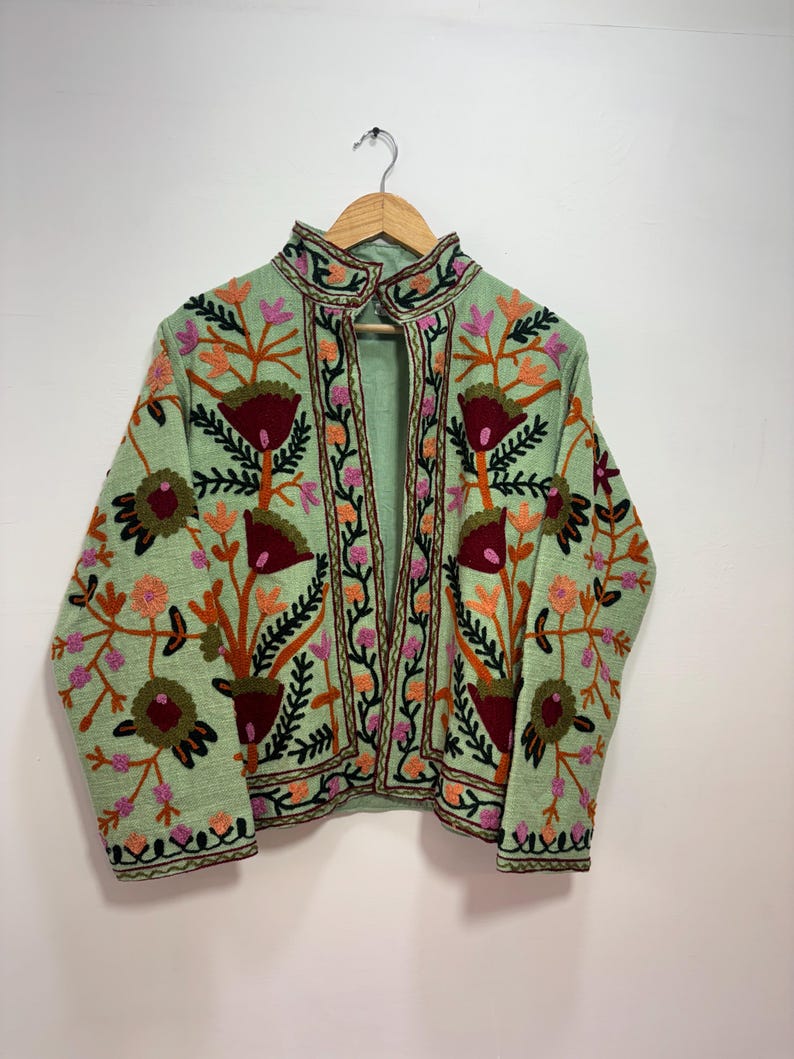 May include: A light green jacket with intricate floral embroidery in shades of red, orange, pink, and green. The jacket features a high collar and is displayed on a wooden hanger. The embroidery creates a textured, decorative pattern.