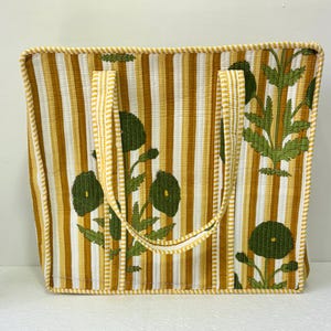 May include: A rectangular tote bag with vertical stripes in shades of yellow and white. The bag features a floral design in green and yellow, with matching handles and trim. The bag is approximately 15 inches wide and 14 inches tall.