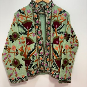 May include: A light green jacket with intricate floral embroidery in shades of red, orange, pink, and black. The jacket features a high collar and is displayed on a wooden hanger. The embroidery creates a detailed pattern across the jacket's surface.
