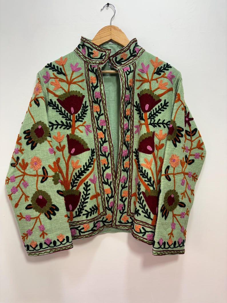 May include: A light green jacket with intricate floral embroidery in shades of red, orange, pink, and black. The jacket features a high collar and is displayed on a wooden hanger. The embroidery creates a detailed pattern across the jacket's surface.