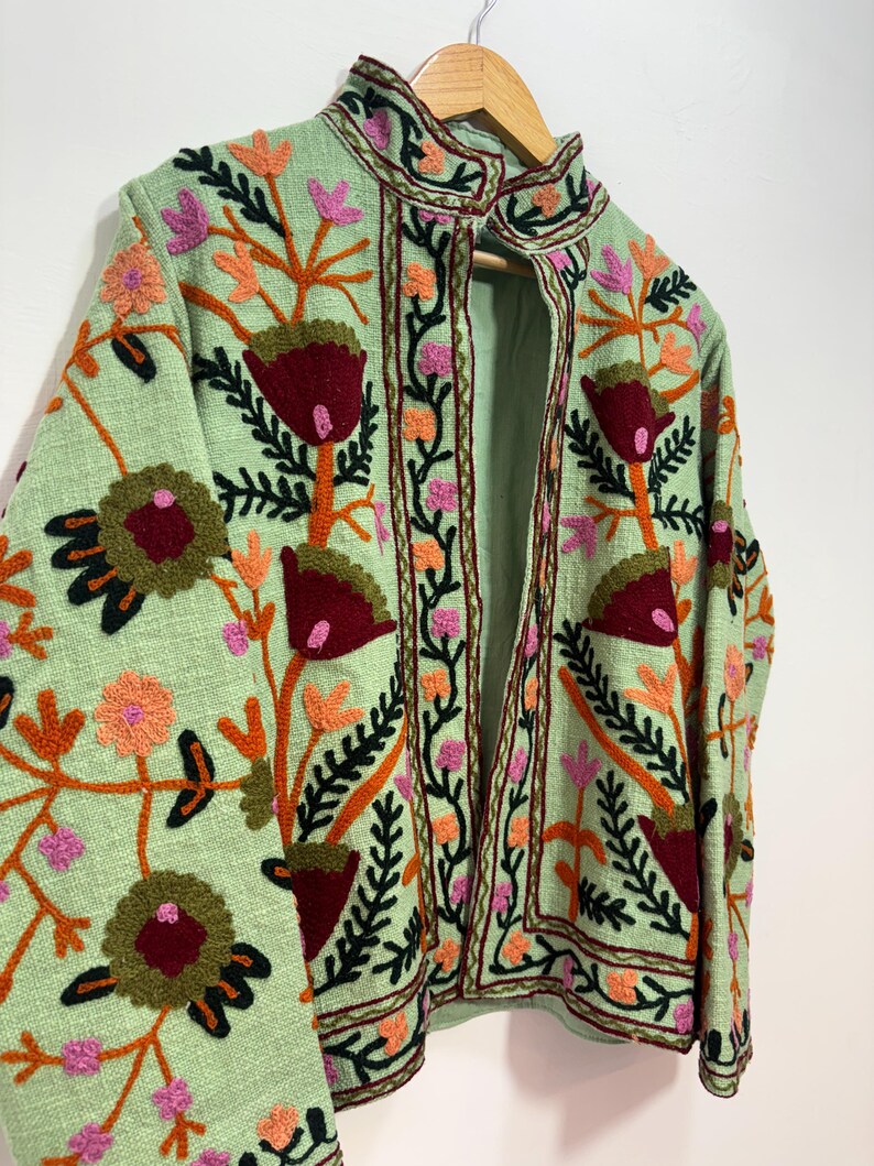 May include: A light green jacket with intricate floral embroidery in shades of red, orange, pink, and green. The jacket features a high collar and is displayed on a wooden hanger. The embroidery creates a textured, artistic design.