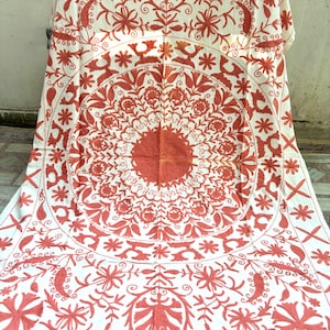 May include: A white cotton tablecloth with a large, intricate red embroidered design. The design features a circular pattern with floral motifs and geometric shapes.