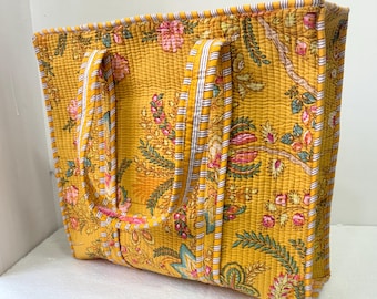 Op de afbeelding: Mosterdgele gewatteerde draagtas met een bloemenpatroon in roze, groen en blauw. De tas heeft twee handvatten met wit en gele strepen. De rechthoekige tas is gemaakt van stof.