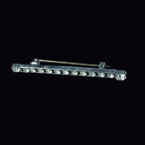 Può includere: Una spilla a barra color argento con una fila orizzontale di piccole pietre chiare sfaccettate incastonate sul davanti. L'oggetto include un sottile meccanismo a spillo in metallo fissato sul retro, progettato per fissare l'accessorio a tessuti o indumenti.