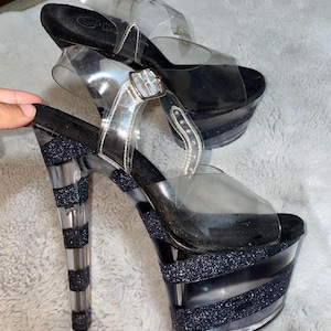 May include: A pair of black and clear platform heels with a glitter design. The heels are clear with black glitter stripes and the straps are clear.