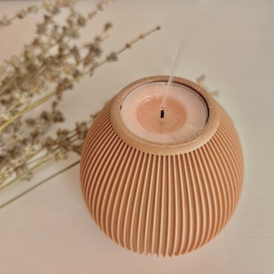 May include: A lit candle in a beige, spherical holder with vertical grooves. The candle is a light pink color and is emitting smoke. Dried lavender sprigs are in the background, creating a calming aesthetic. The candle holder is a decorative item.