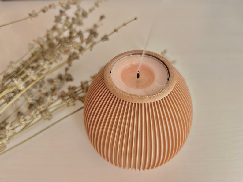 May include: A lit candle in a beige, spherical holder with vertical grooves. The candle is a light pink color and is emitting smoke. Dried lavender sprigs are in the background, creating a calming aesthetic. The candle holder is a decorative item.