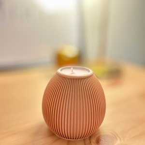 May include: A peach-colored, ribbed candle in a round, egg-like holder. The candle has a white wick and sits on a wooden surface. The holder has vertical grooves, creating a textured appearance. The background is blurred.