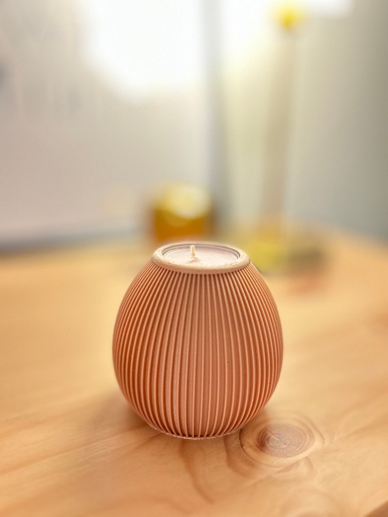 May include: A peach-colored, ribbed candle in a round, egg-like holder. The candle has a white wick and sits on a wooden surface. The holder has vertical grooves, creating a textured appearance. The background is blurred.