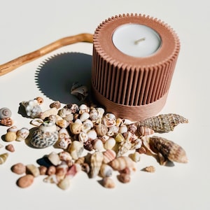 May include: A brown cylindrical candle holder with a white candle inside. The holder is surrounded by a scattering of small seashells.