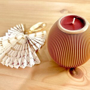 May include: A decorative angel made of folded paper with a floral pattern and a gold ribbon. Next to it is a wooden candle holder with a red candle inside. The candle holder has vertical grooves.