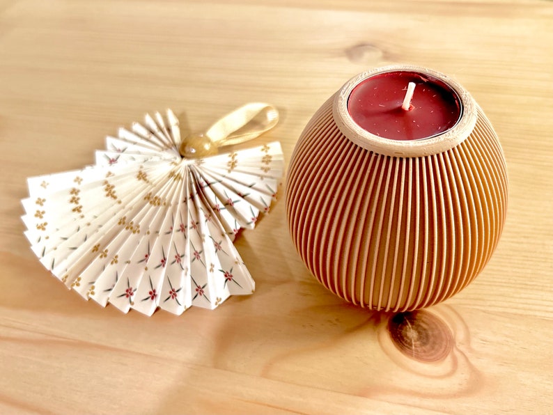 May include: A decorative angel made of folded paper with a floral pattern and a gold ribbon. Next to it is a wooden candle holder with a red candle inside. The candle holder has vertical grooves.