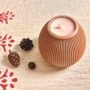 May include: A decorative candle holder with a pink candle inside. The holder is a light brown color with vertical lines. The candle holder is sitting on a white surface with red floral patterns. There are three pinecones next to the candle holder.