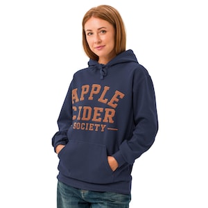 May include: A navy blue hooded sweatshirt featuring a front kangaroo pocket and the text APPLE CIDER SOCIETY printed in a textured orange font across the chest. The hoodie includes matching navy blue drawstrings at the neckline.