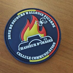 Peut inclure: Un écusson rond noir avec du texte blanc qui dit "Zone de Secours Wallonie Picarde" et "Cellule Communication". L'écusson présente également un appareil photo blanc avec une flamme rouge et jaune derrière et un drone blanc au-dessus de la flamme.